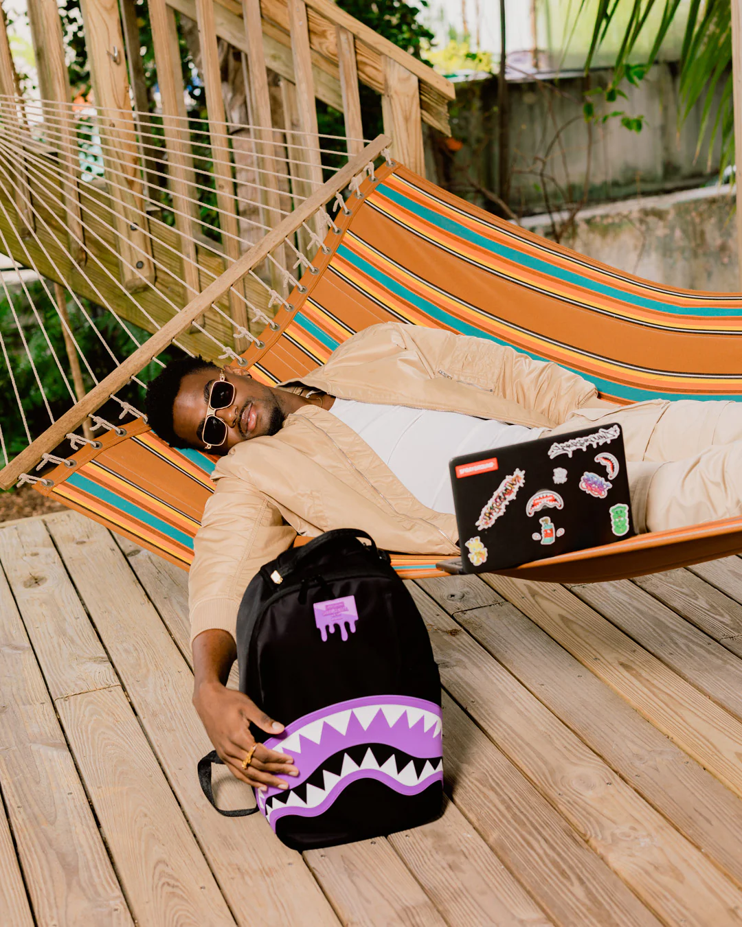 Person lying in a hammock with a laptop and backpack, holding a backpack with a shark design.
