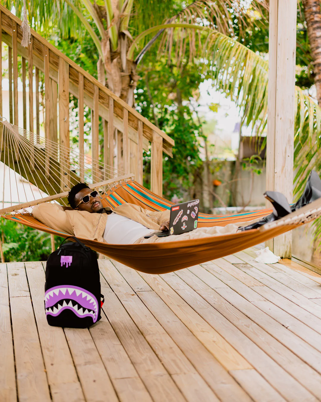 Person lying in a hammock on a wooden deck with a laptop, surrounded by greenery.