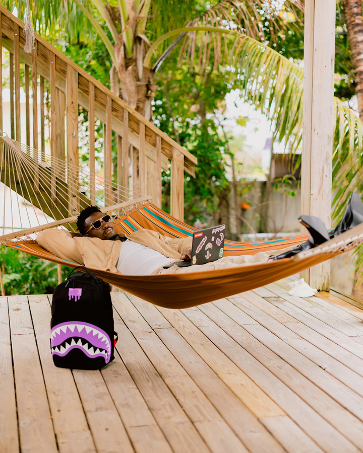 Person lying in a hammock on a wooden deck with a laptop, surrounded by greenery.