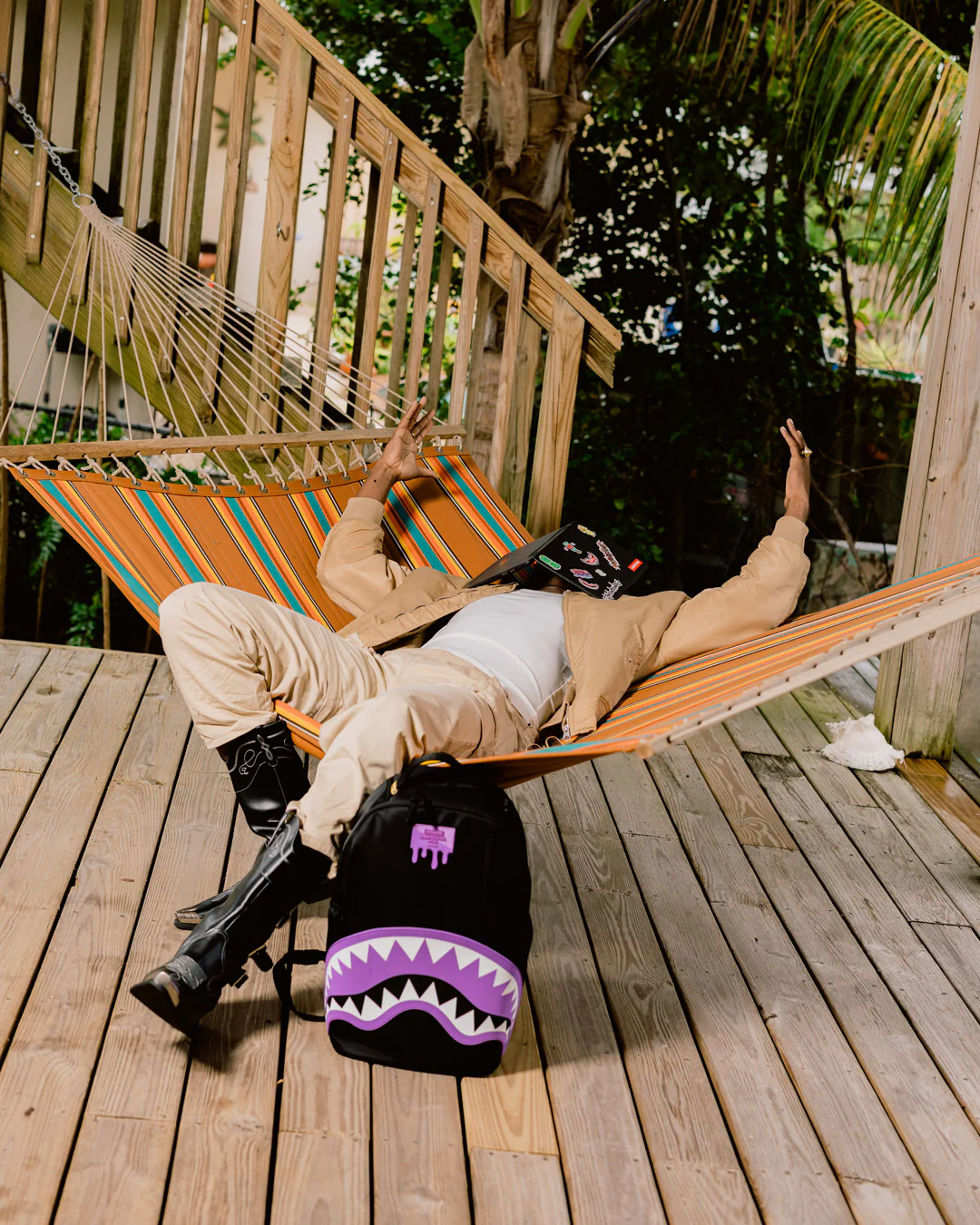 Person lounging in a striped hammock on a wooden deck with a purple backpack featuring a shark design.