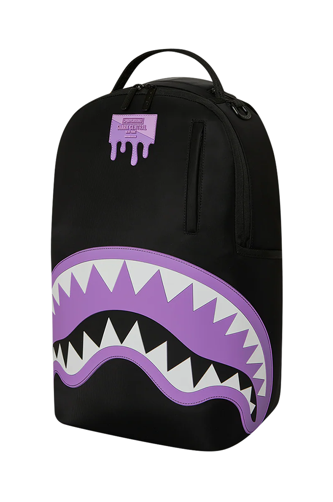A black backpack featuring a large purple and white shark mouth graphic on the front, with a small purple Sprayground logo patch near the top.