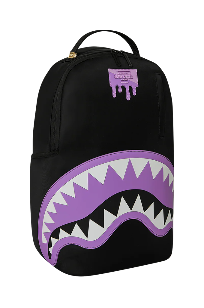 A black backpack featuring a large purple and white shark mouth graphic on the front, with a small purple Sprayground logo patch near the top.