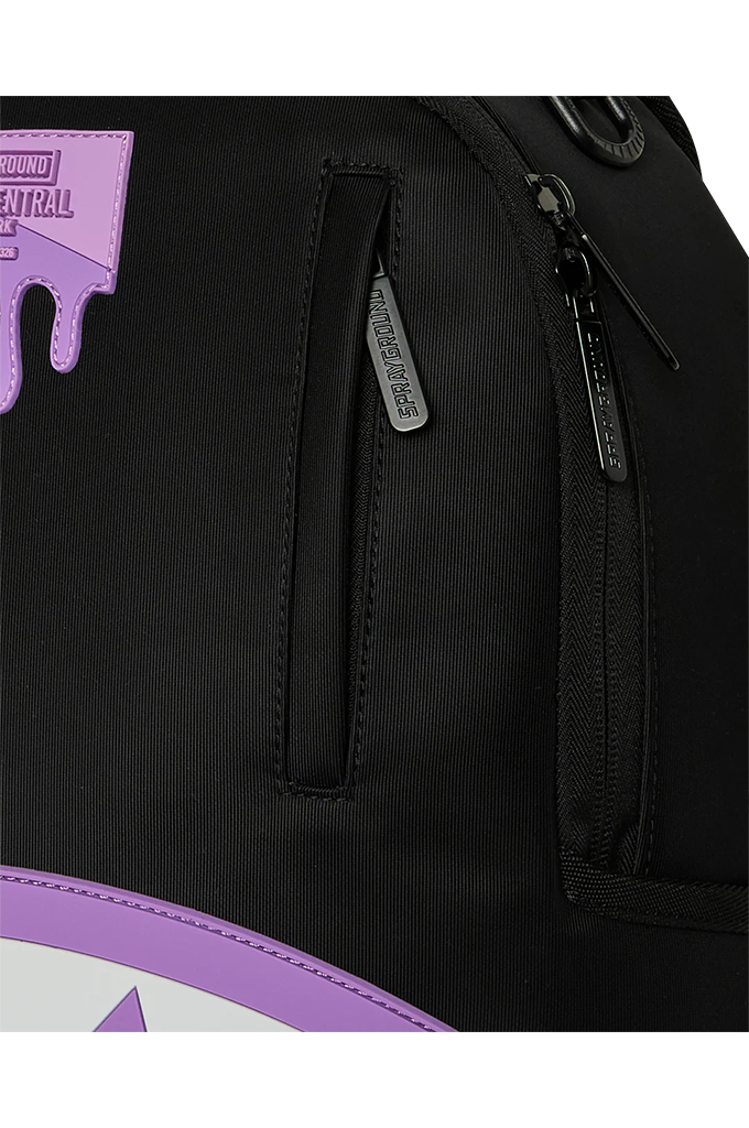 Close-up of a black backpack showing a vertical zippered pocket with a silver-tone zipper pull labeled 'SPRAYGROUND', a side zipper, and a purple logo patch on the upper left.