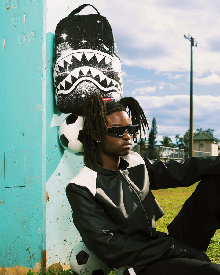 Person sitting outdoors with a black and white backpack and soccer ball, wearing sunglasses and a black and white jacket.