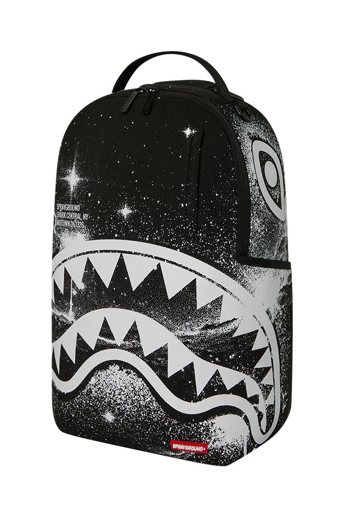 A black backpack featuring a white shark face graphic and a starry galaxy print, with a small red Sprayground logo tag at the bottom.