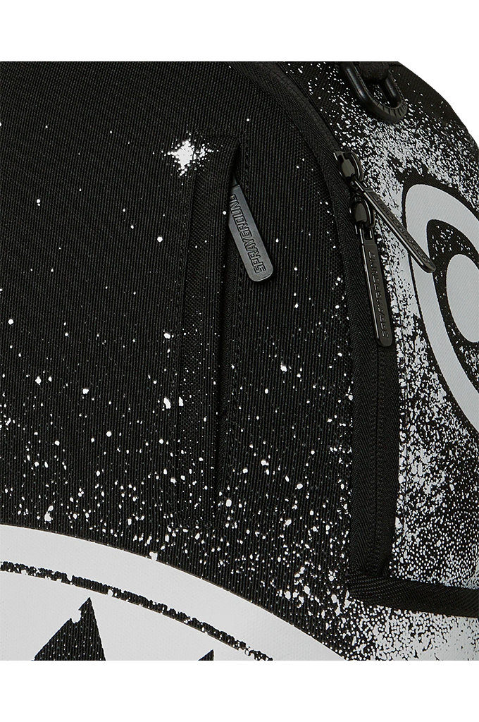 Close-up of a black backpack with a white speckled galaxy print, featuring a side pocket with a gray zipper pull labeled 'SPRAYGROUND' and a large white graphic on the side.