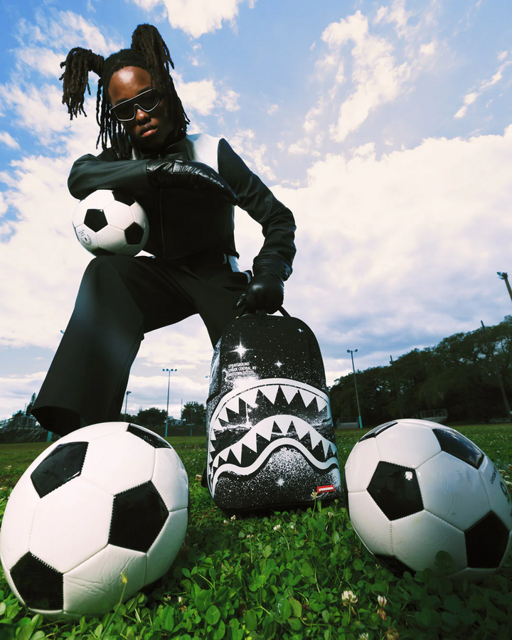 Person sitting on a grassy field with soccer balls and a shark-patterned bag.