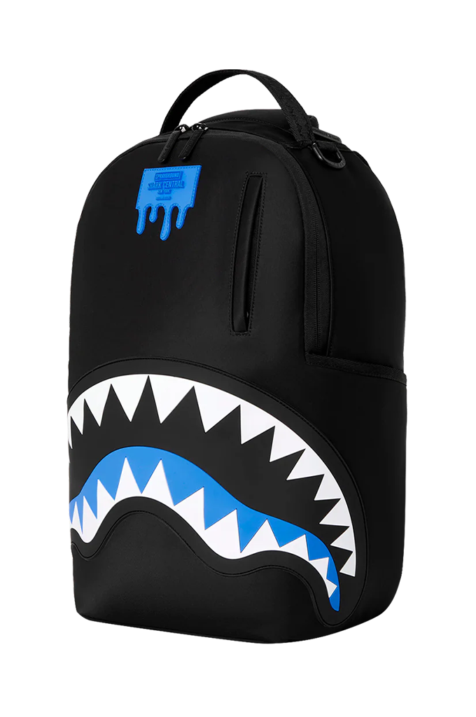 A black Sprayground backpack featuring the iconic shark mouth graphic with white and blue details, and a blue Spray-R logo patch on the upper front.