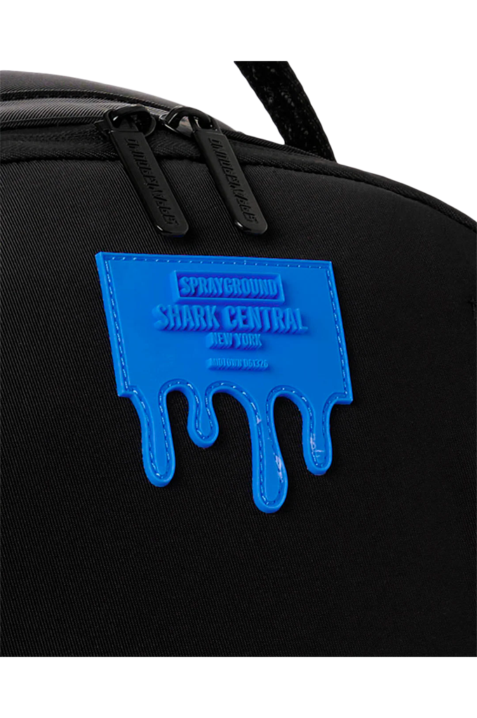 Close-up of a black backpack featuring a bright blue dripping patch with the text 'SPRAYGROUND SHARK CENTRAL NEW YORK' and black zipper pulls with the same branding.