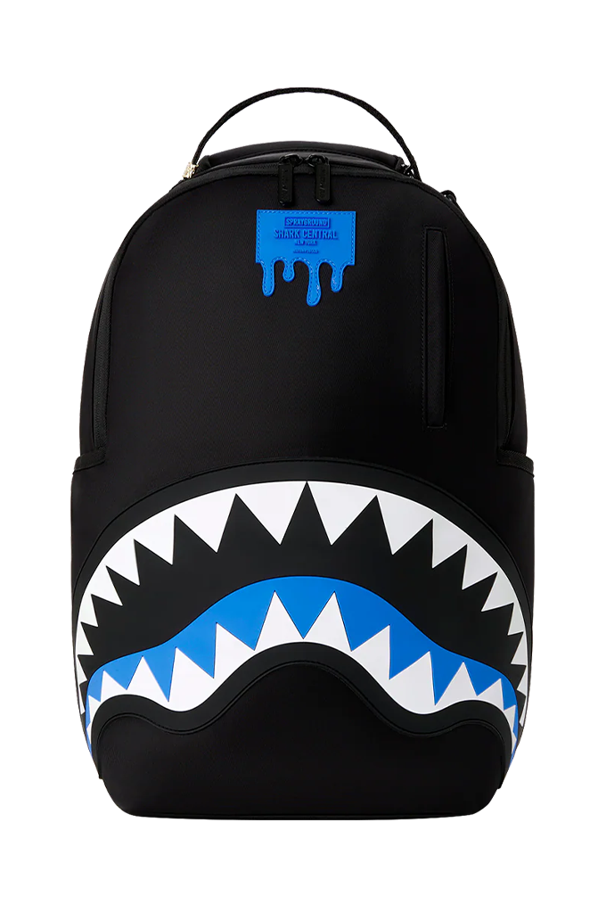 A black Sprayground backpack featuring the iconic shark mouth graphic with white and blue details, and a blue Spray-R logo patch on the upper front.