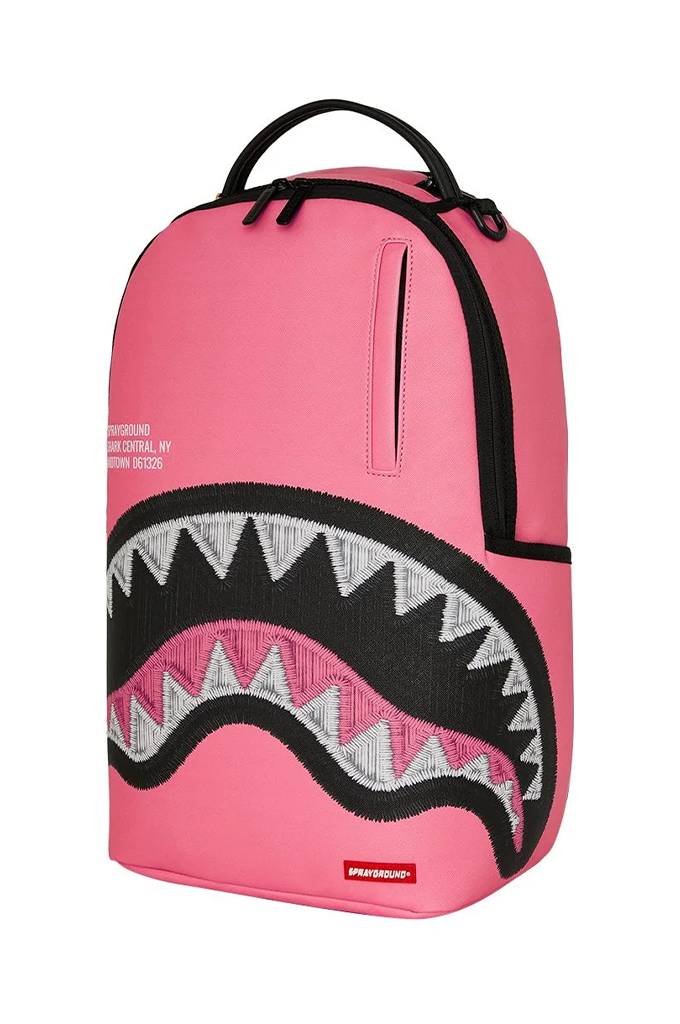 A pink Sprayground Shark backpack with a large embroidered shark mouth design on the front, featuring black, white, and pink details, a top handle, and a zippered pocket.