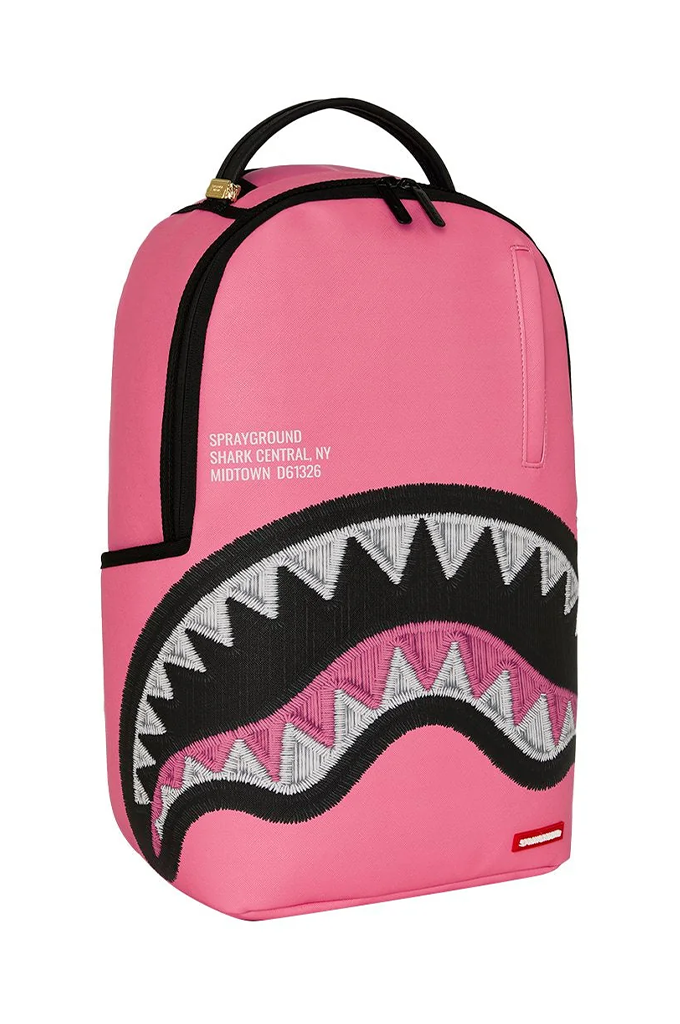 A pink Sprayground Shark backpack with a large embroidered shark mouth design in black, white, and pink on the front, featuring a top carry handle and zippered compartments.