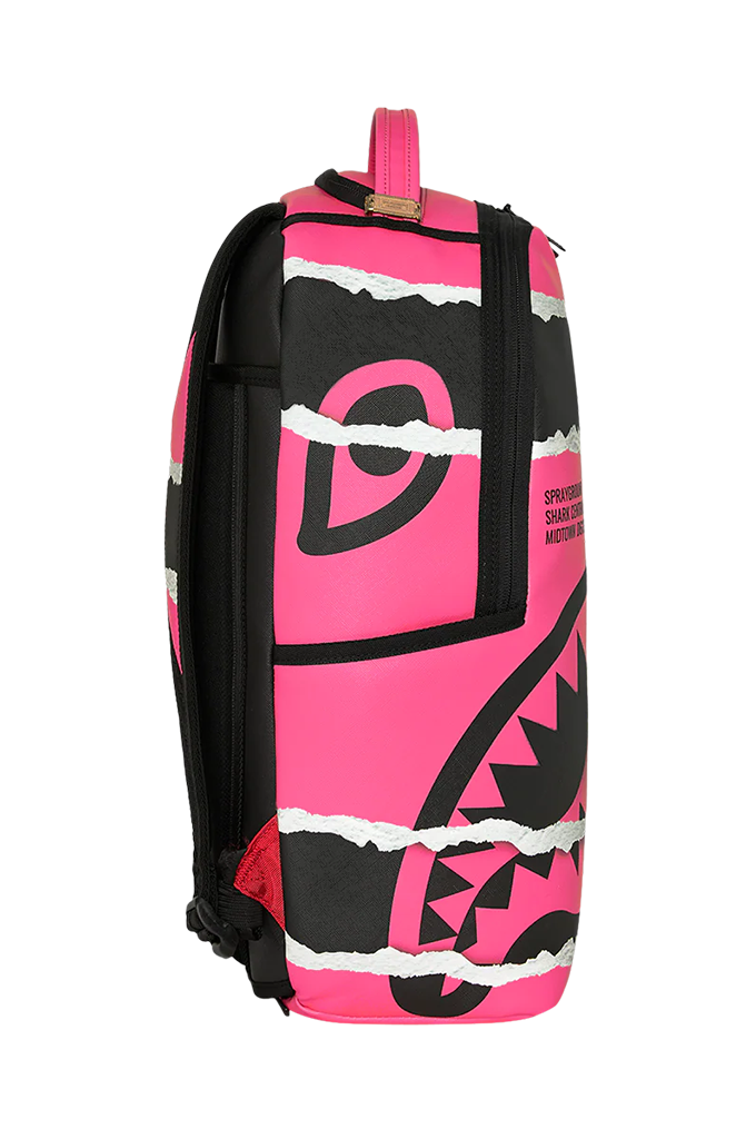 A pink and black backpack with a torn-paper graphic design, featuring a shark mouth motif and the brand name 'Sprayground' printed on the side.