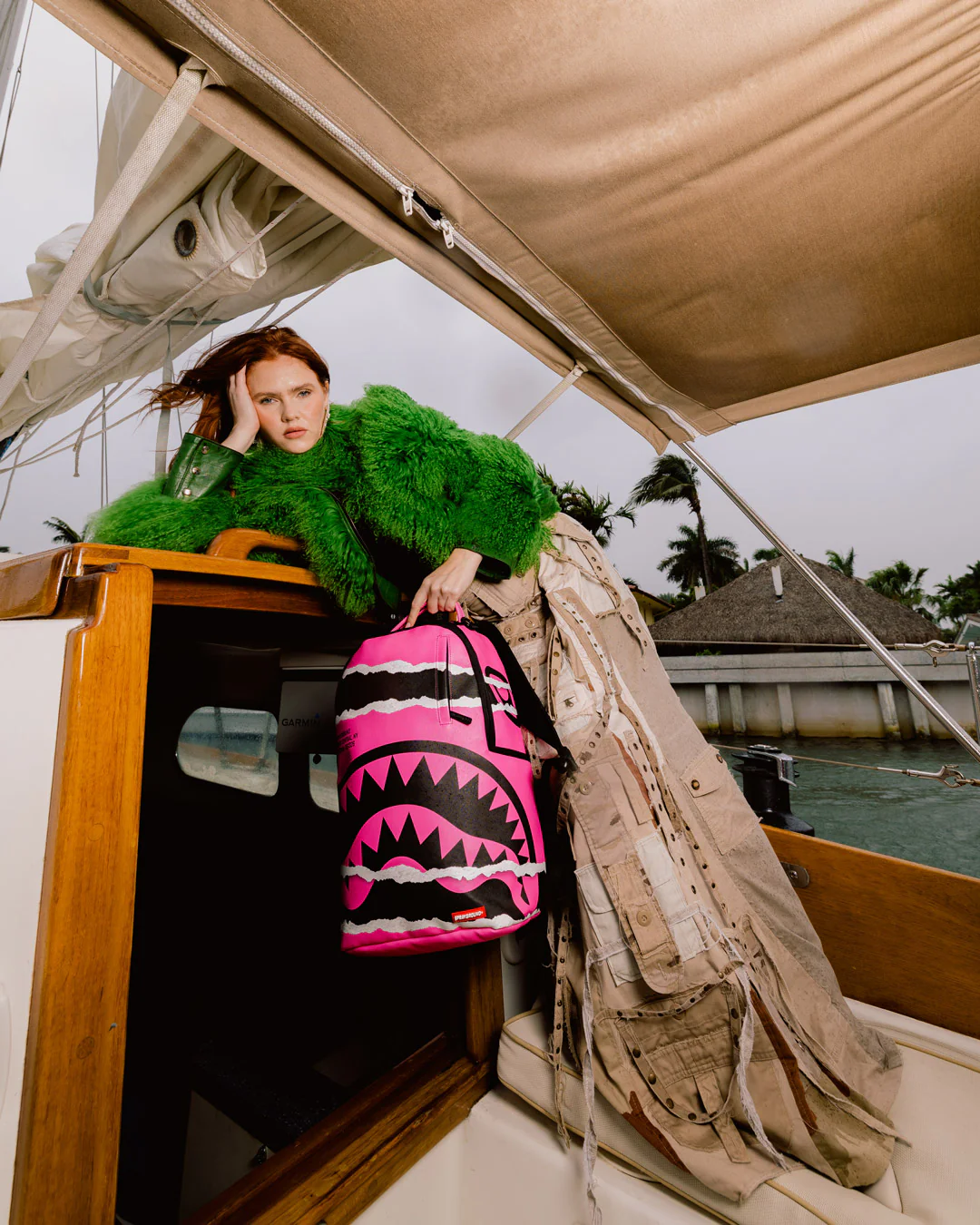 A model wearing a bright green fuzzy coat and a beige distressed jacket leans over the edge of a boat cabin, holding a pink and black shark-mouth backpack. The background shows water, distant buildings, and palm trees under an overcast sky.