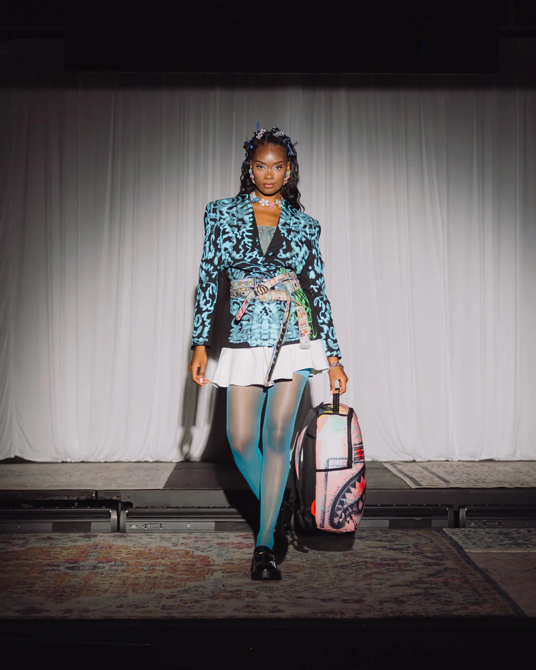 Model walking on a runway wearing a colorful outfit with a patterned jacket and white skirt, holding a suitcase.