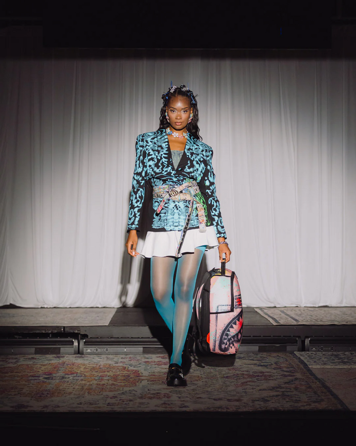 Model walking on a runway wearing a colorful outfit with a patterned jacket and white skirt, holding a suitcase.