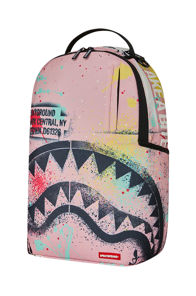 A pink Sprayground backpack featuring a large black shark mouth graphic, colorful paint splatters, and a black top handle, shown on a plain white background.