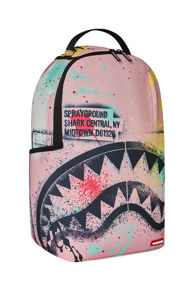 A Sprayground Shark backpack in pink with a shark mouth graphic, paint splatters, and a zippered pocket on the front.