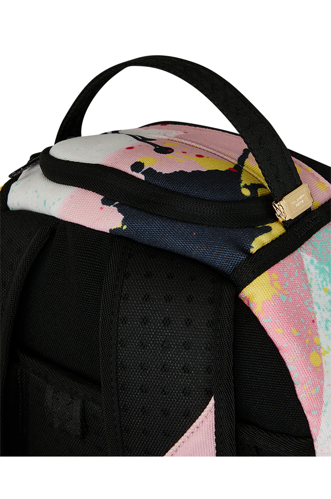 Close-up of a backpack top corner showing a black top handle, black padded shoulder strap, zipper trim, and a multicolor paint-splatter pattern in pink, yellow, blue, and white.