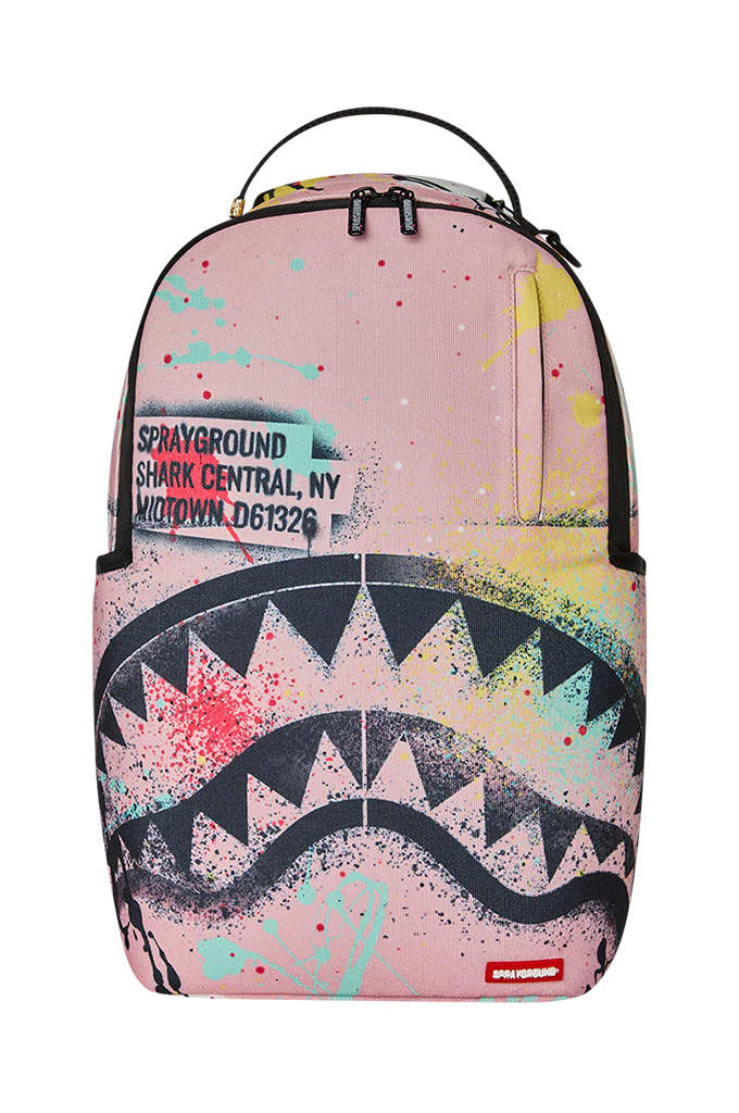 A Sprayground Shark backpack in pink with a black shark mouth graphic, colorful paint splatters, and a zippered pocket on the front.