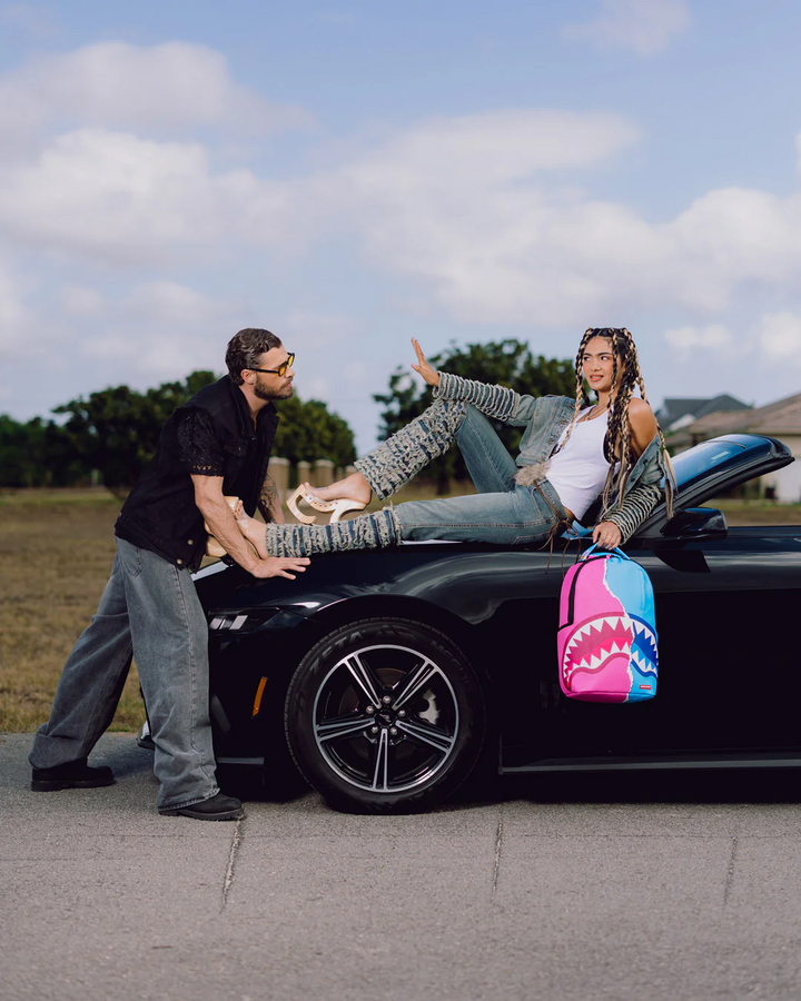 Two people with a black car and a colorful shark-patterned bag on a road.