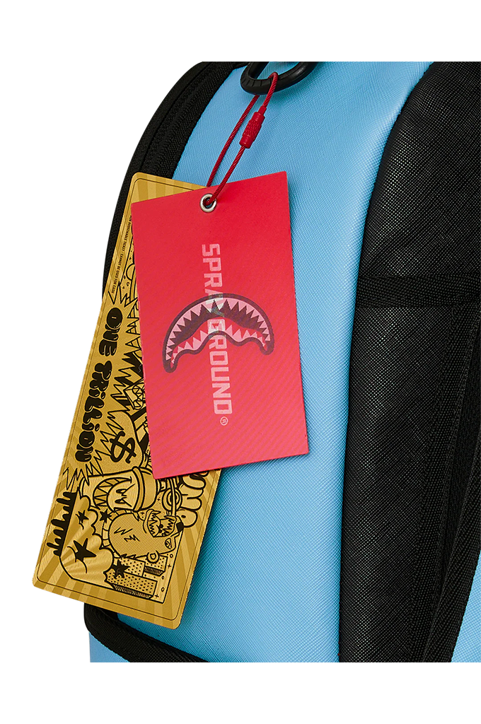A close-up of a light blue and black backpack with a red hangtag featuring a shark mouth graphic and the text 'SPRAYGROUND', alongside a yellow graphic luggage tag.