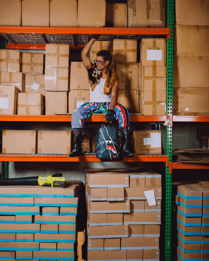A person wearing a white tank top, colorful patterned pants, and black boots sits on an orange warehouse shelf surrounded by stacked cardboard boxes, holding a black backpack with a shark graphic.