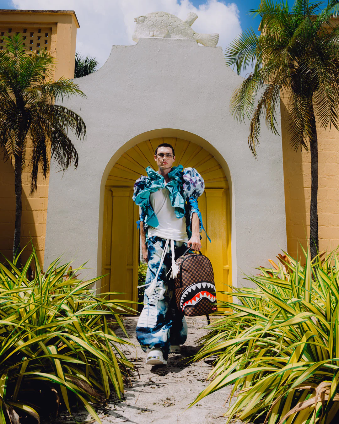 A man walks down a sandy path in front of a white stucco archway with yellow doors, wearing a bright blue patterned jacket, matching blue patterned pants, and carrying a brown patterned backpack with a shark-face graphic.