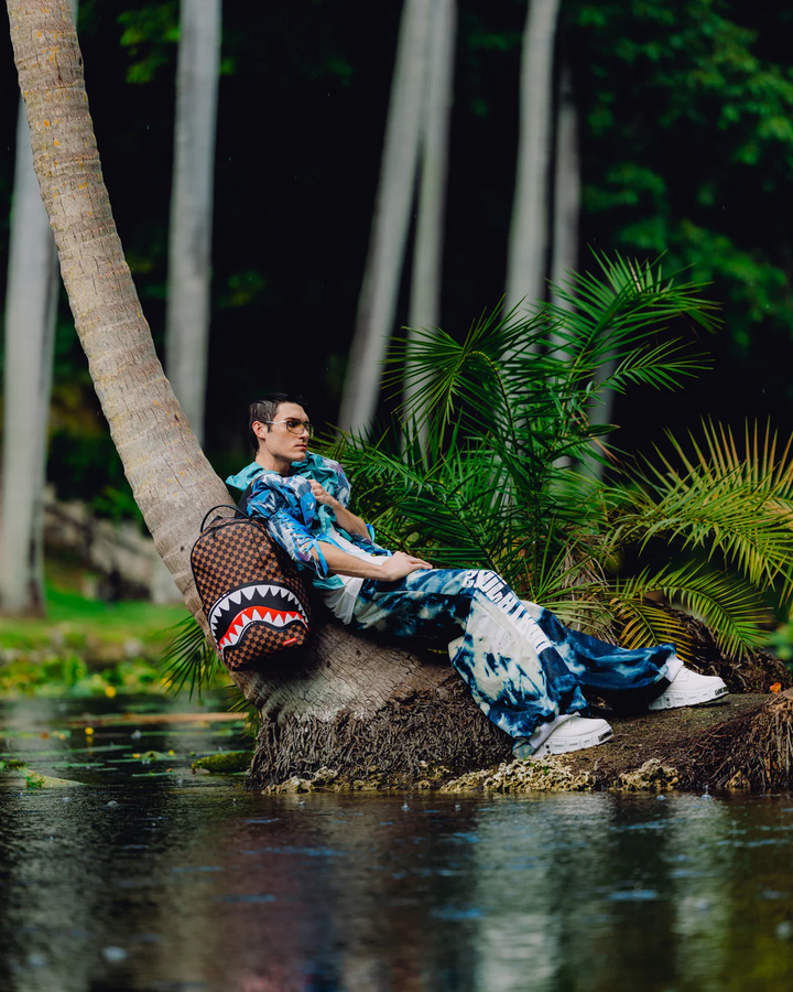 A model wearing a blue and white patterned shirt and matching pants sits on a tree trunk in a tropical outdoor setting with palm trees and water, carrying a brown woven shoulder bag with a shark mouth design.