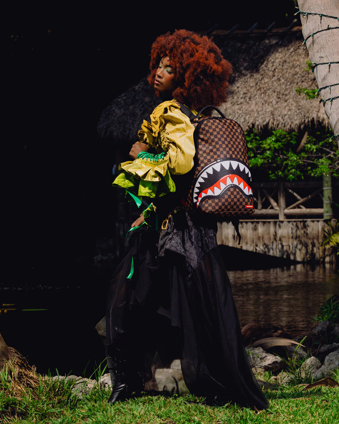 A model with a large curly red-brown afro stands outdoors near water and a thatched structure, wearing a black dress and carrying a brown checkered backpack with a shark-face graphic.