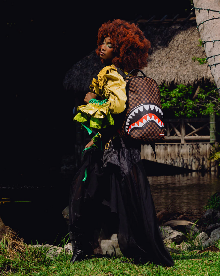 A model with a large curly red-brown afro stands outdoors near water and a thatched structure, wearing a black dress and carrying a brown checkered backpack with a shark-face graphic.