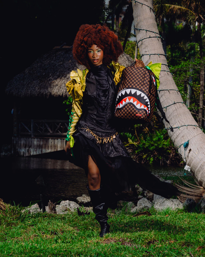 A model wearing a black ruffled dress, gold and green jacket, and black boots stands outdoors near palm trees and a thatched structure, carrying a brown shark-mouth backpack.