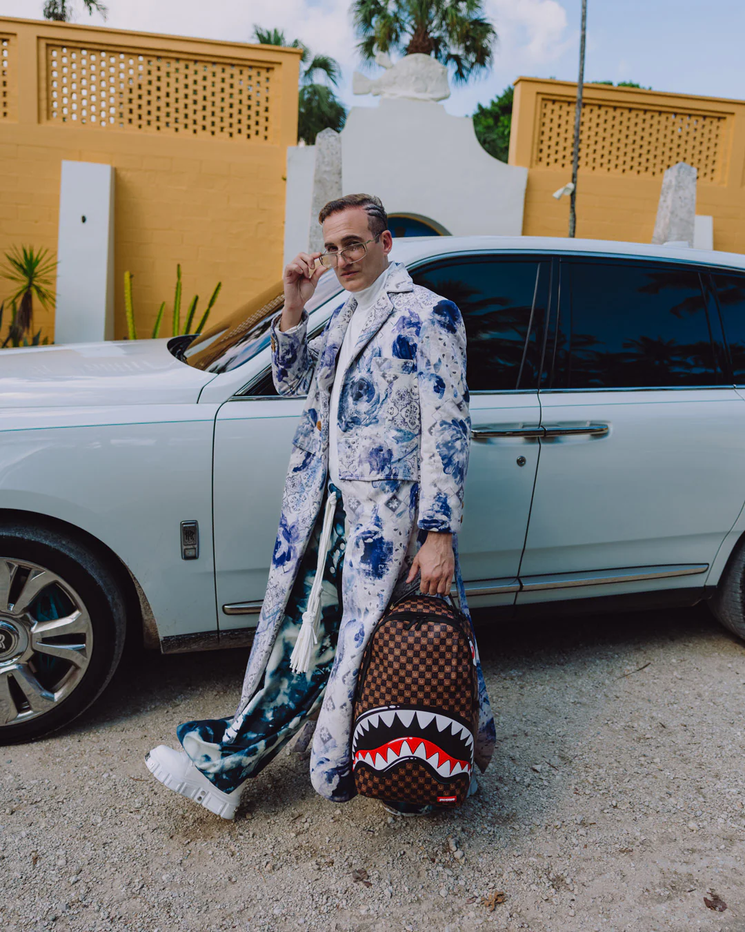 A man wearing a long white coat with a blue and green floral print stands next to a white Rolls-Royce, holding a brown monogram backpack with a shark-mouth graphic.