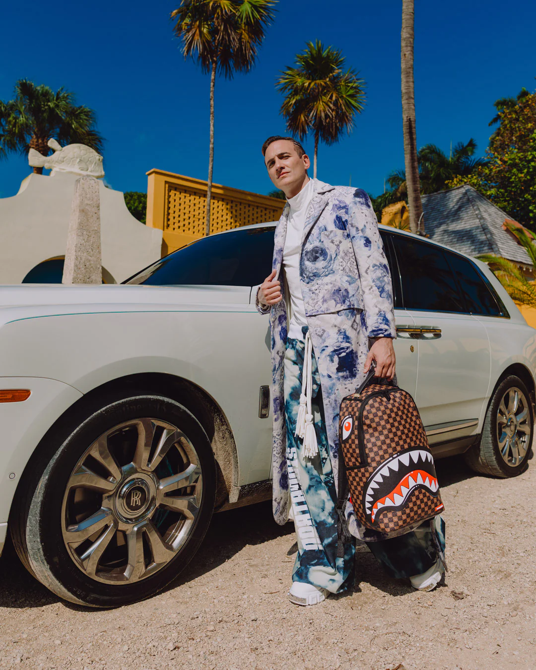 A man wearing a blue and white patterned suit stands next to a white Rolls-Royce car in a sunny outdoor setting with palm trees, holding a brown checkered backpack with a shark-mouth graphic.