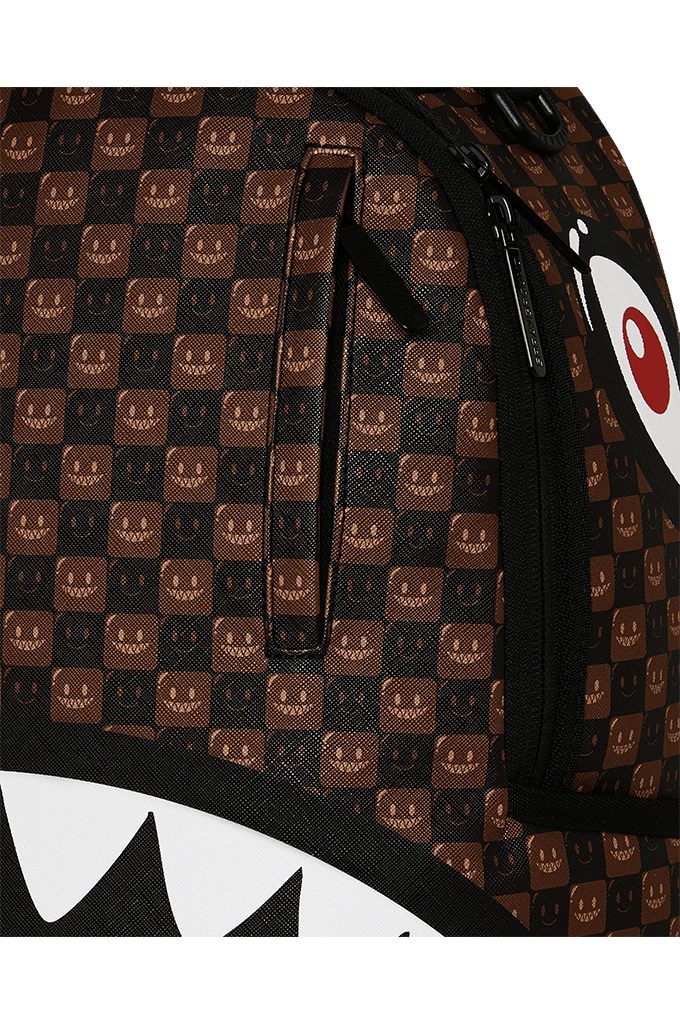 Close-up of a brown and black patterned backpack featuring a repeating shark-face motif and a partial white-and-red eye graphic, with black zippers and trim.