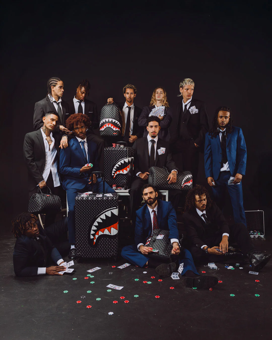 A group of men in suits and blue suits pose with multiple black Shark Tank-style suitcases decorated with a white shark mouth graphic, set against a dark studio background with scattered poker chips on the floor.
