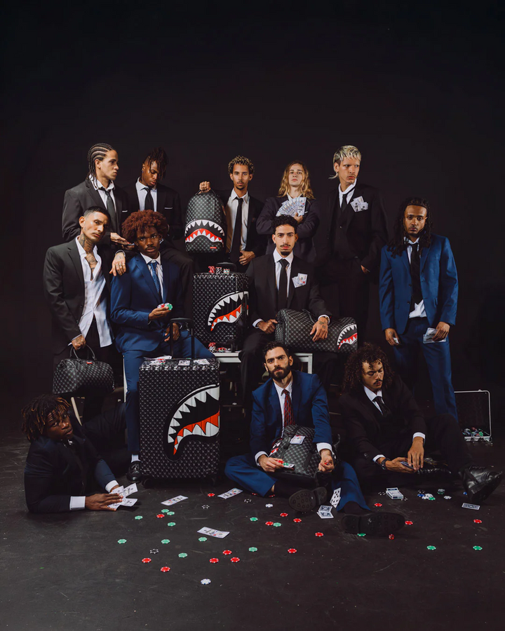 A group of men in suits and blue suits pose with multiple black Shark Tank-style suitcases decorated with a white shark mouth graphic, set against a dark studio background with scattered poker chips on the floor.