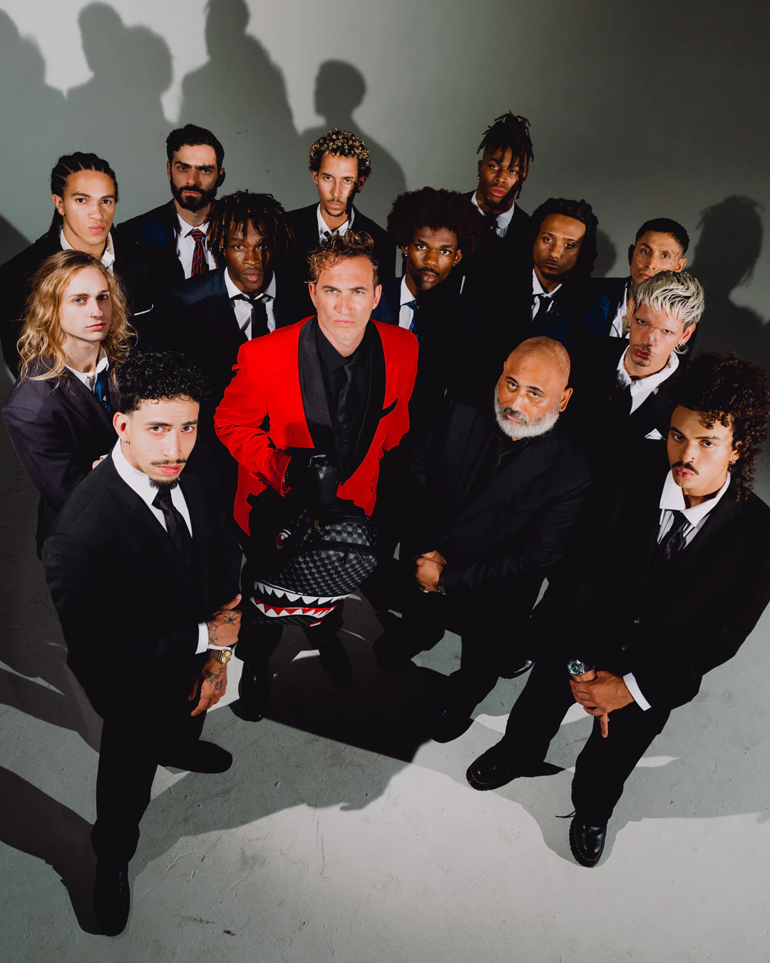 A group of men in formal suits and ties pose together in a studio setting. One man in the center wears a bright red blazer over a black outfit, while most others wear black suits.