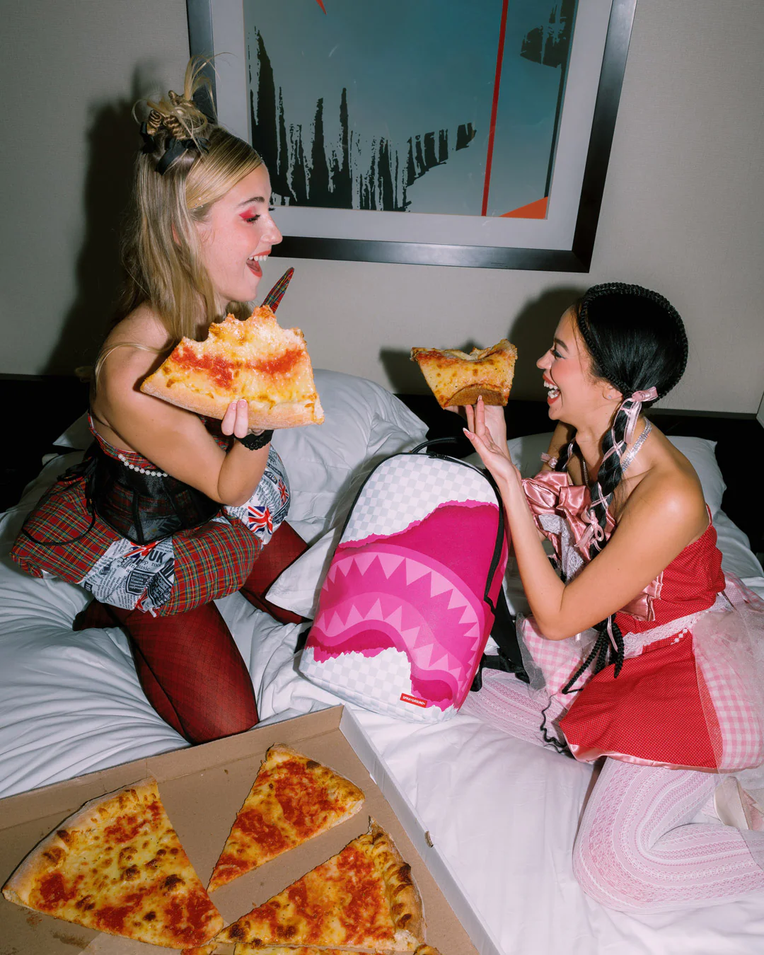 Two women sitting on a bed eating pizza slices, with a large pink and white shark-mouth backpack in the center and an open pizza box in the foreground.