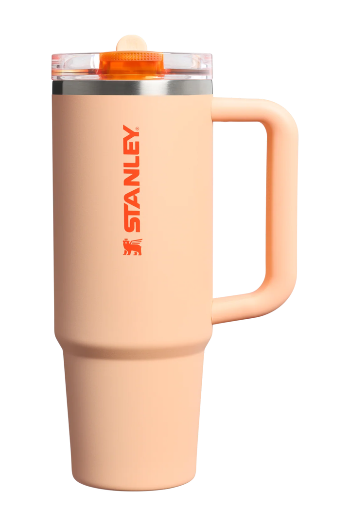 A Stanley travel mug in a peachy pink color with a matching handle, a clear lid, and an orange accent on the lid and the vertical STANLEY logo.#color_orange-sherbet