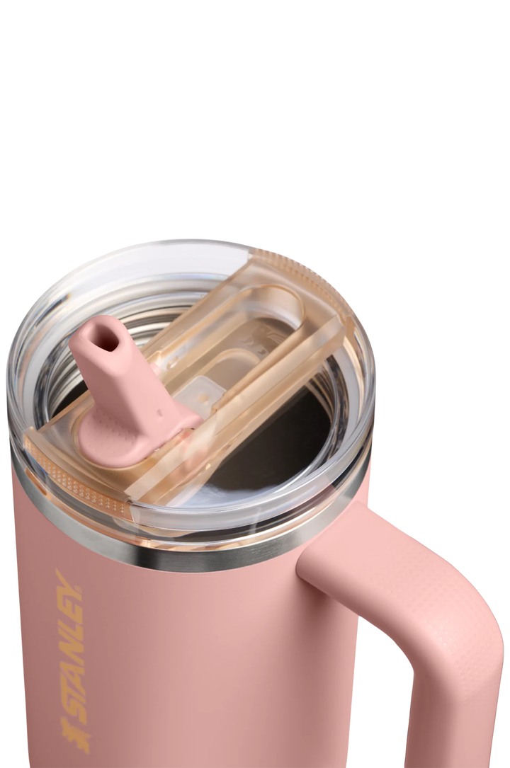 Close-up of a pink Stanley insulated tumbler with a clear lid, gold-toned rim, and a pink straw spout, shown on a white background.#color_peach-rose