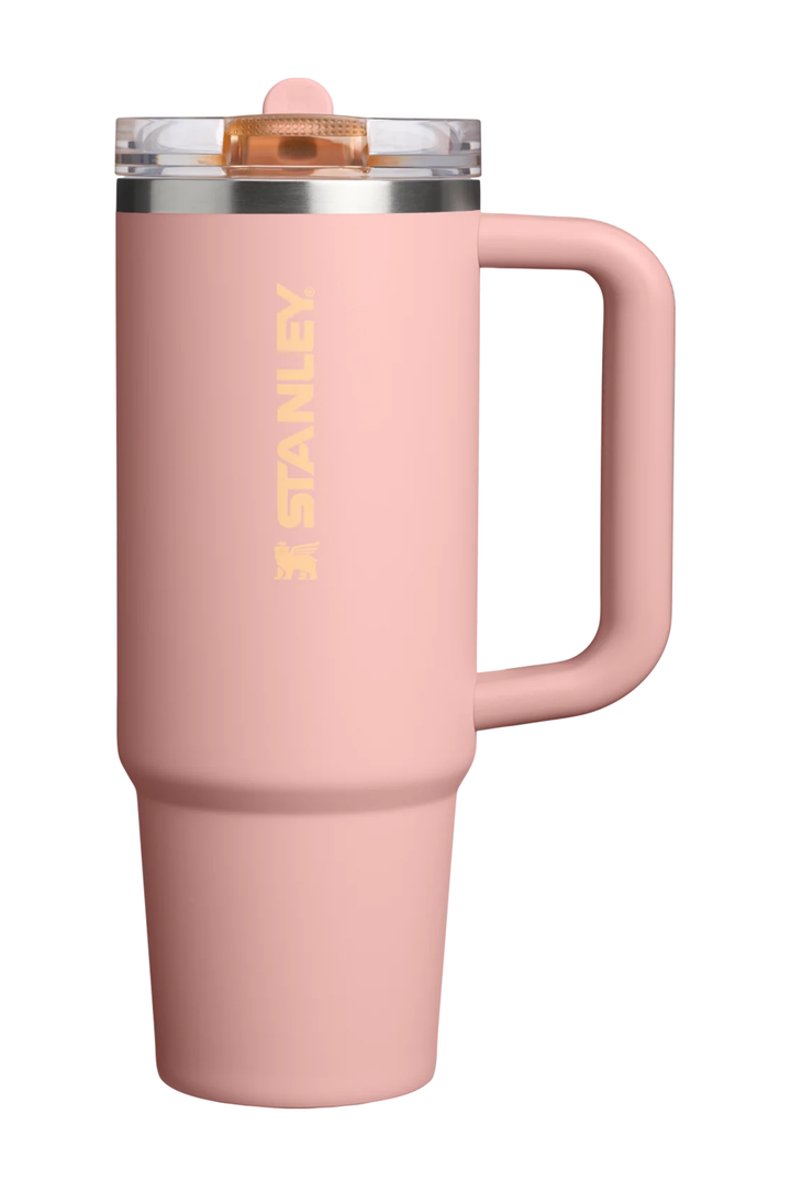 A pink Stanley travel mug with a handle, clear lid, and gold Stanley logo on a white background.#color_peach-rose