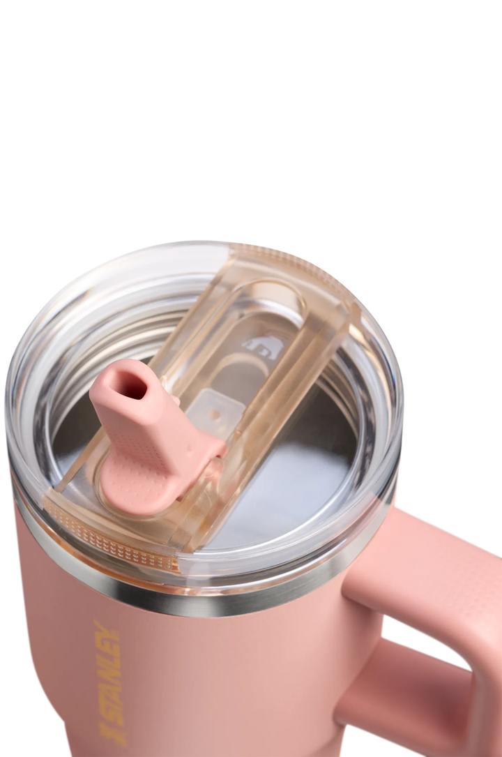 Close-up of a pink insulated tumbler with a clear lid and pink straw, showing part of the handle and a gold-toned vertical logo on a white background.#color_peach-rose