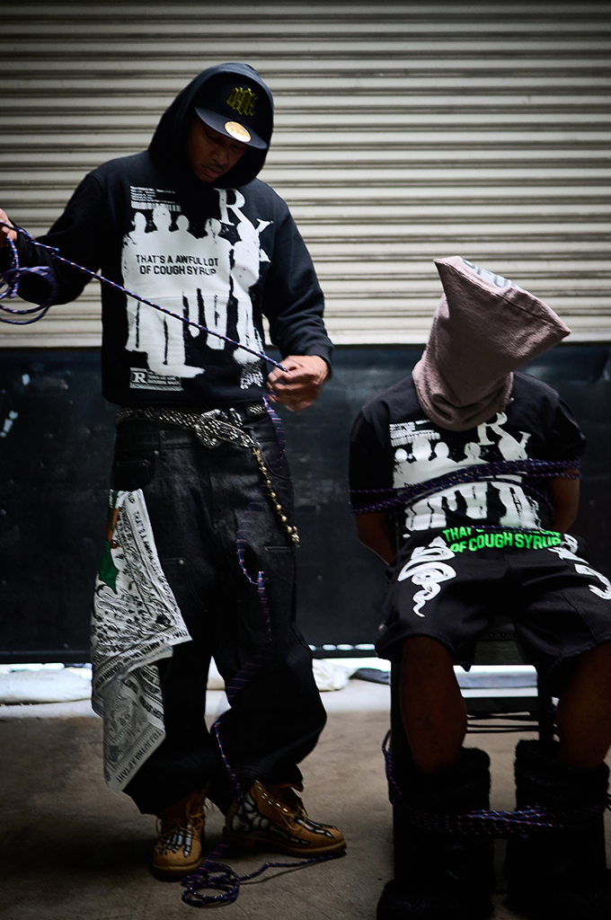 A model stands wearing a black hooded sweatshirt from the brand "That's A Awful Lot Of Cough Syrup." The hoodie features a large white graphic of silhouetted figures alongside bold text reading "THAT'S A AWFUL LOT OF COUGH SYRUP." The model is wearing a black baseball cap under the hood, distressed black wide-leg jeans with a patterned bandana hanging from the side pocket, and a multi-layered metallic waist chain. 