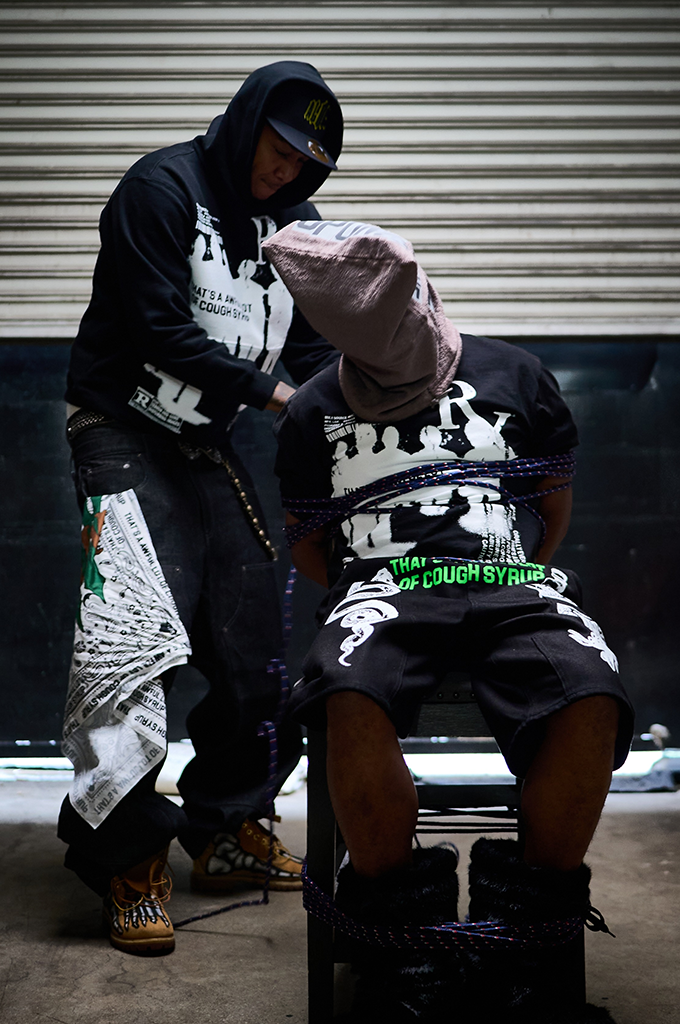 Streetwear model wearing a black "That's An Awful Lot of Cough Syrup" Big Pharma pullover hoodie and graphic pants next to a seated model in a matching graphic t-shirt and shorts.