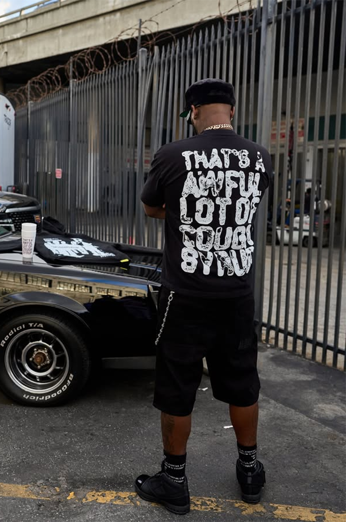 A man standing outdoors from a back view wearing a black streetwear short-sleeve graphic t-shirt featuring the bold, textured text "THAT'S A AWFUL LOT OF COUGH SYRUP" printed in distressed white lettering across the upper back.