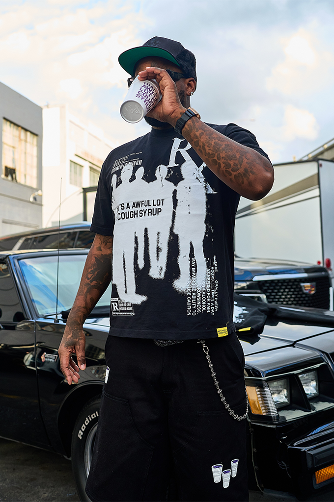 A man wearing the black ALOCS Pharma Tee from the brand That's A Awful Lot Of Cough Syrup leans against a black classic car. The graphic t-shirt features a prominent white Rx symbol and human silhouettes on the front. He styles the streetwear look with matching black graphic sweatshorts, a black wallet chain, and a dark baseball cap while holding a white double cup.