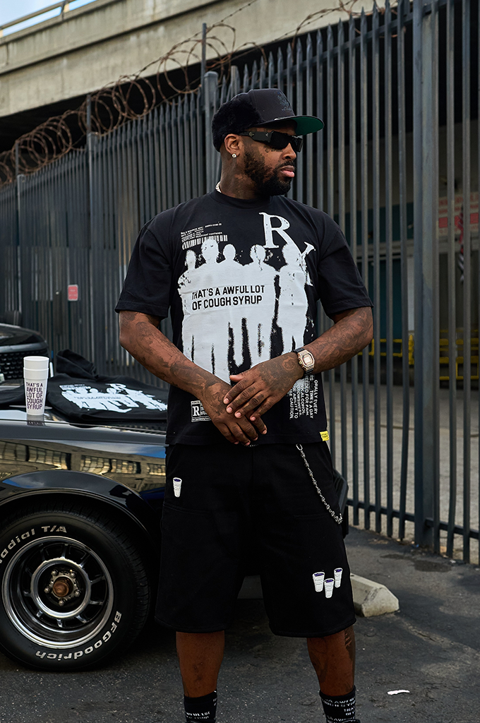 A man wearing the black That's A Awful Lot Of Cough Syrup ALOCS Pharma graphic tee and matching logo shorts, standing next to a black car.