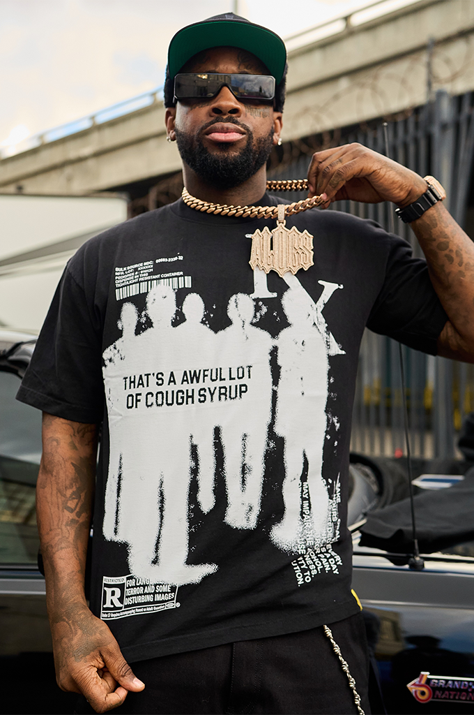 A man wearing a black streetwear graphic t-shirt, a chunky gold chain with an "ALOCS" wordmark pendant, sunglasses, and a green trucker hat. The t-shirt features a bold white screen-printed graphic on the front showing three silhouettes with a vertical barcode design and text that reads "THAT'S A AWFUL LOT OF COUGH SYRUP." An "R" rating warning block is printed on the lower right corner of the graphic. The man is posing outdoors in front of a metal fence, adjusting his sunglasses with one hand.