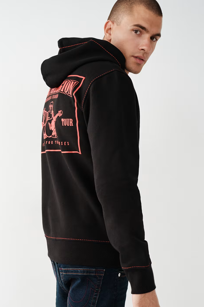 A man wearing a black hoodie with red contrast stitching and a red graphic print on the back, paired with dark blue jeans, photographed against a light gray studio background.